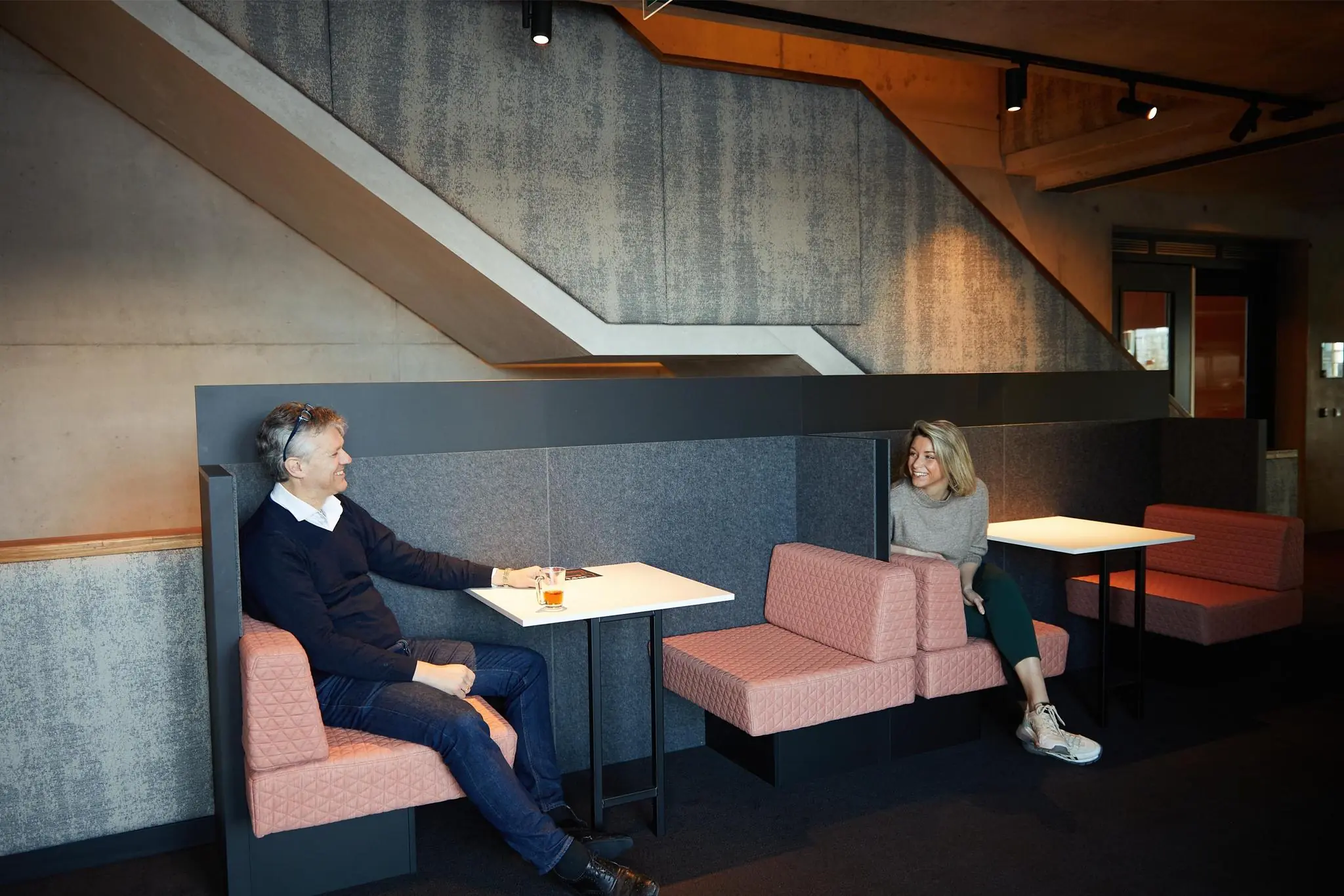 Twee mensen zitten tegenover elkaar in een moderne zithoek onder een betonnen trap in een kantoor aan de Orteliuslaan.