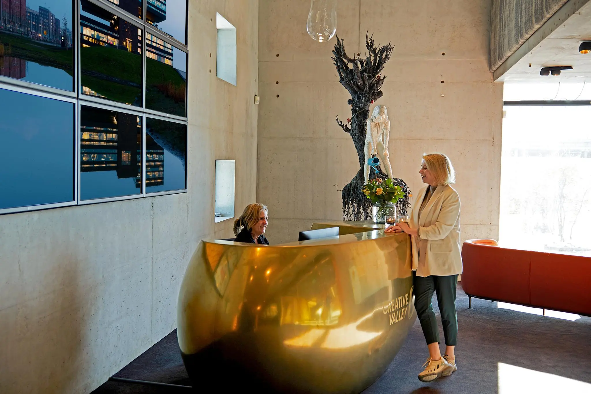 Vrouw staat bij een gouden receptiebalie van Creative Valley aan de Orteliuslaan, met kunstwerk en reflecterende wandpanelen op de achtergrond.