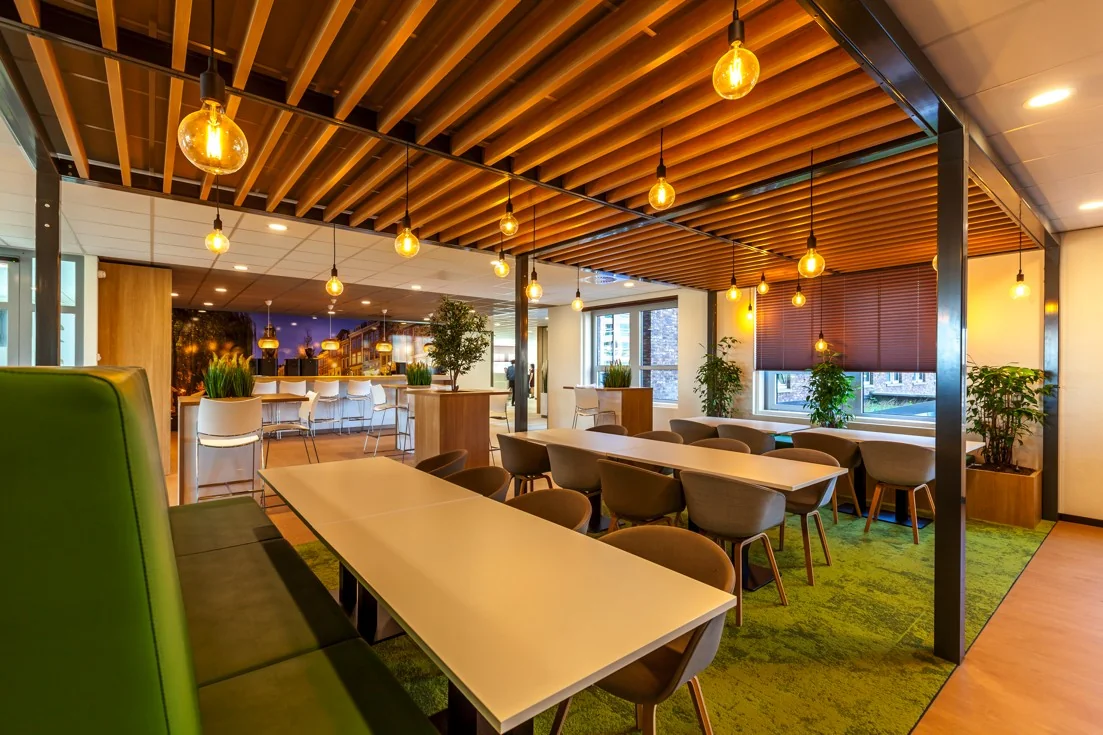 Moderne kantoorkantine aan de Orteliuslaan met houten plafondaccenten, hanglampen en groene vloerbedekking.
