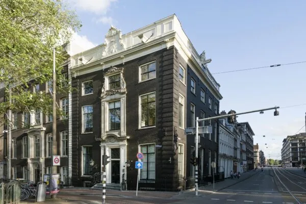 Hoekpand aan de Herengracht in Amsterdam met een klassieke gevel en witte ornamenten, gelegen aan een kruispunt met verkeerslichten.