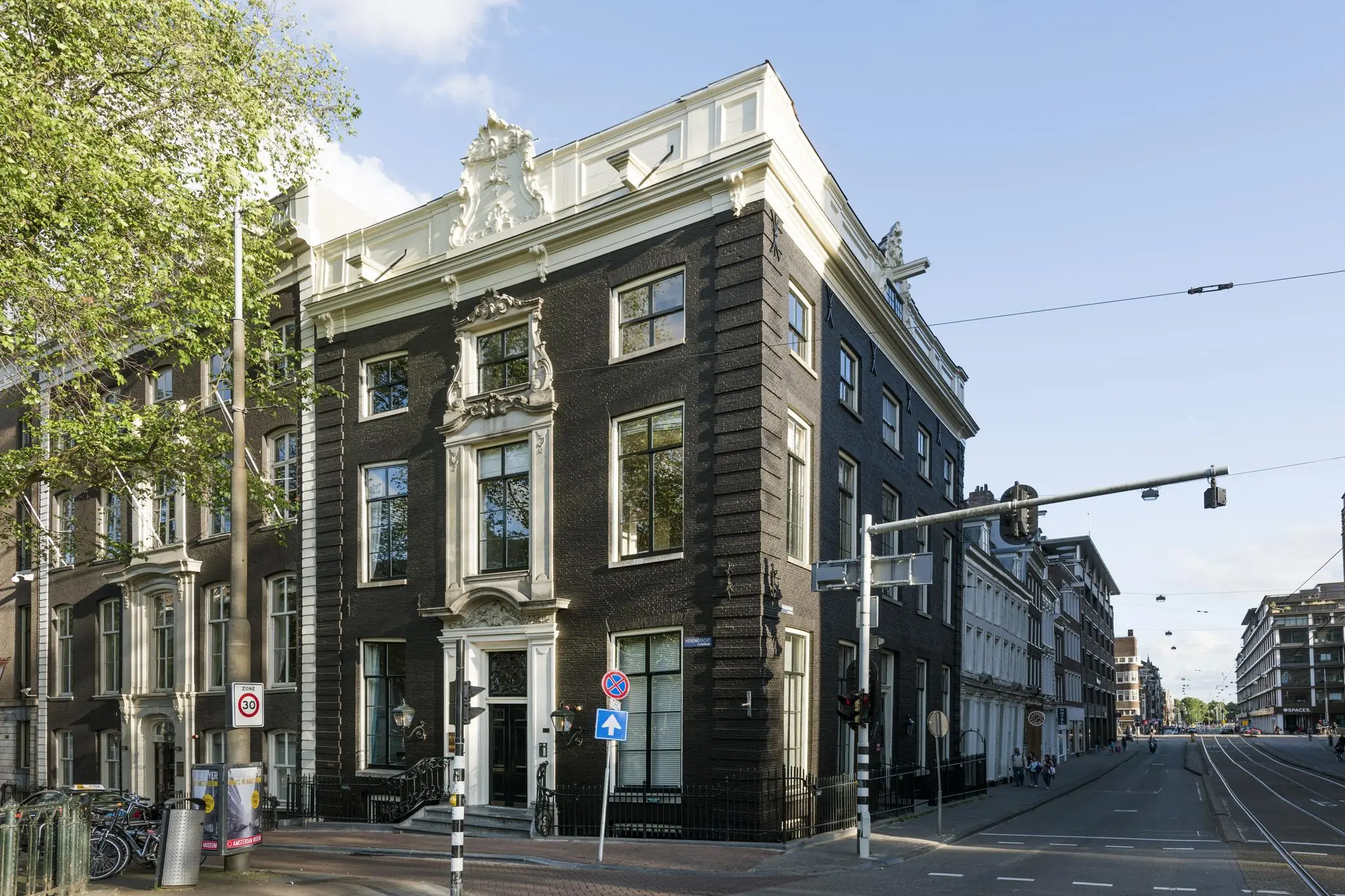 Hoekpand aan de Herengracht in Amsterdam met een klassieke gevel en witte ornamenten, gelegen aan een kruispunt met verkeerslichten.