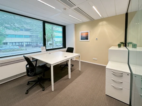 Een moderne, lichte kantoorruimte met twee bureaus, stoelen, en een groot raam met uitzicht naar buiten.