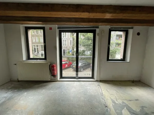 Uitzicht vanuit een lege kamer op het Entrepotdok met drie ramen, een brandblusser en zicht op geparkeerde auto's buiten.