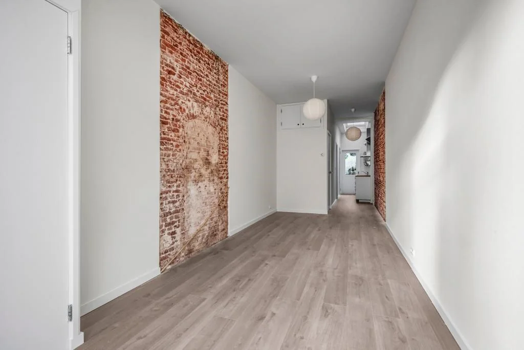 Langwerpige, smalle woonkamer met een houten vloer, witte muren en twee decoratieve bakstenen muurdelen aan weerszijden.