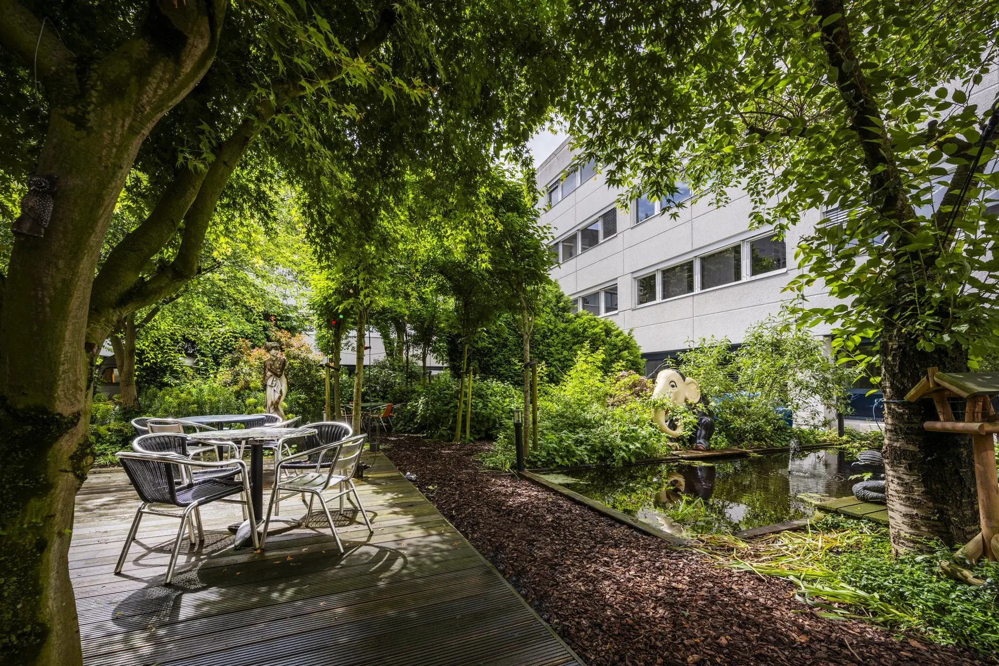 Groene binnentuin bij Diemermere met zitgedeelte, kunstobjecten en een vijver naast een kantoorgebouw.