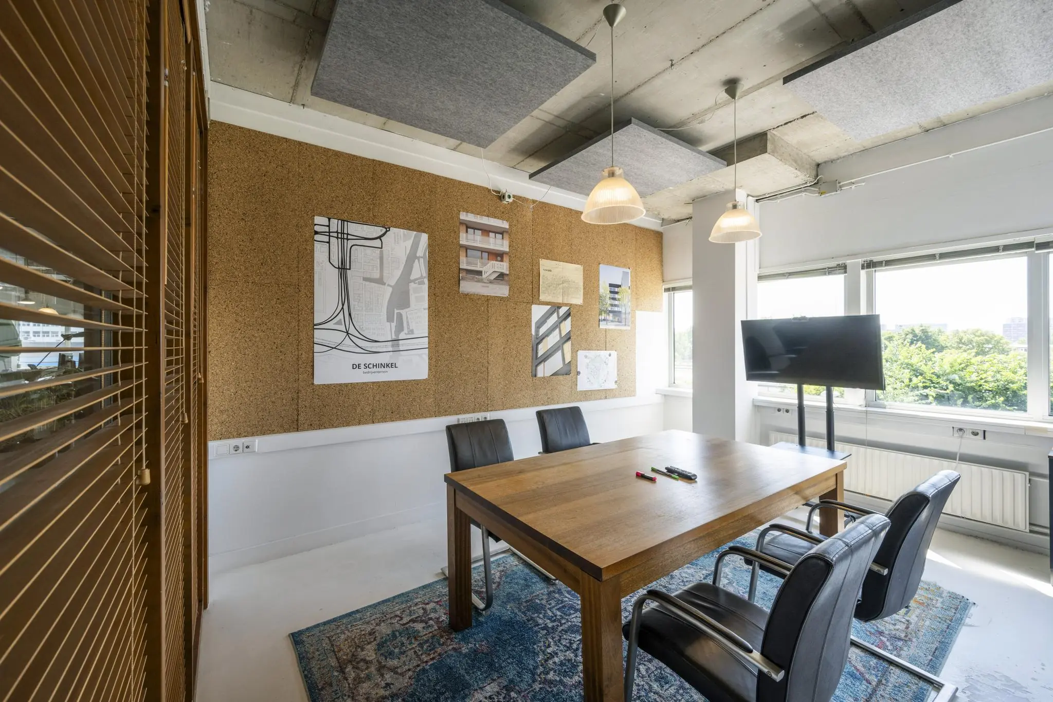 Vergaderruimte met houten tafel, vier stoelen, een prikbord met projectposters en een tv-scherm bij het raam aan de Anthony Fokkerweg.