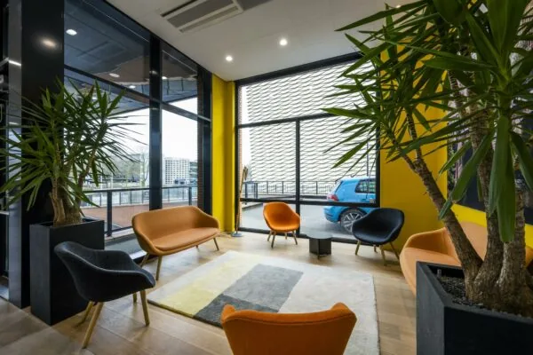 Moderne wachtruimte aan de Winthontlaan met kleurrijke stoelen, grote planten en uitzicht op een blauwe auto buiten.