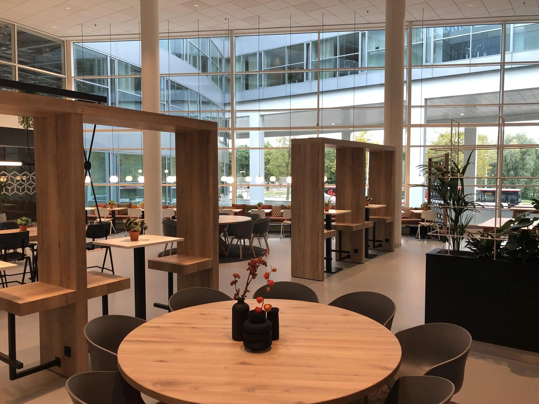 Moderne en open kantoorkantine aan de Newtonlaan met houten tafels, zithoeken en grote ramen met uitzicht op buiten.