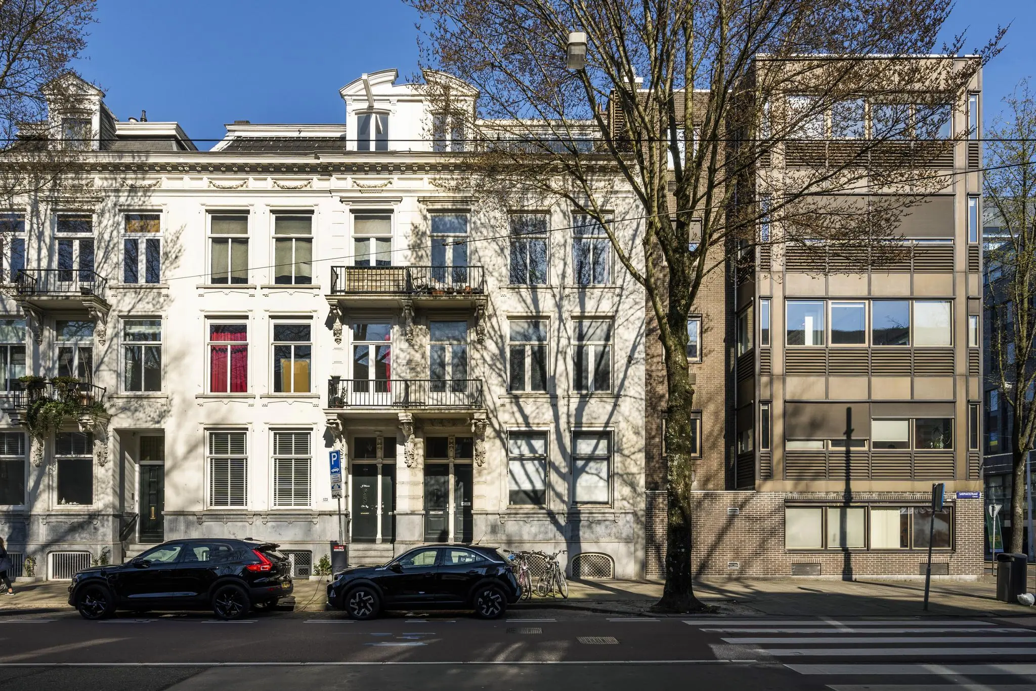 Voorgevel van historische en moderne gebouwen aan de Sarphatistraat in Amsterdam met geparkeerde auto's en fietsen.