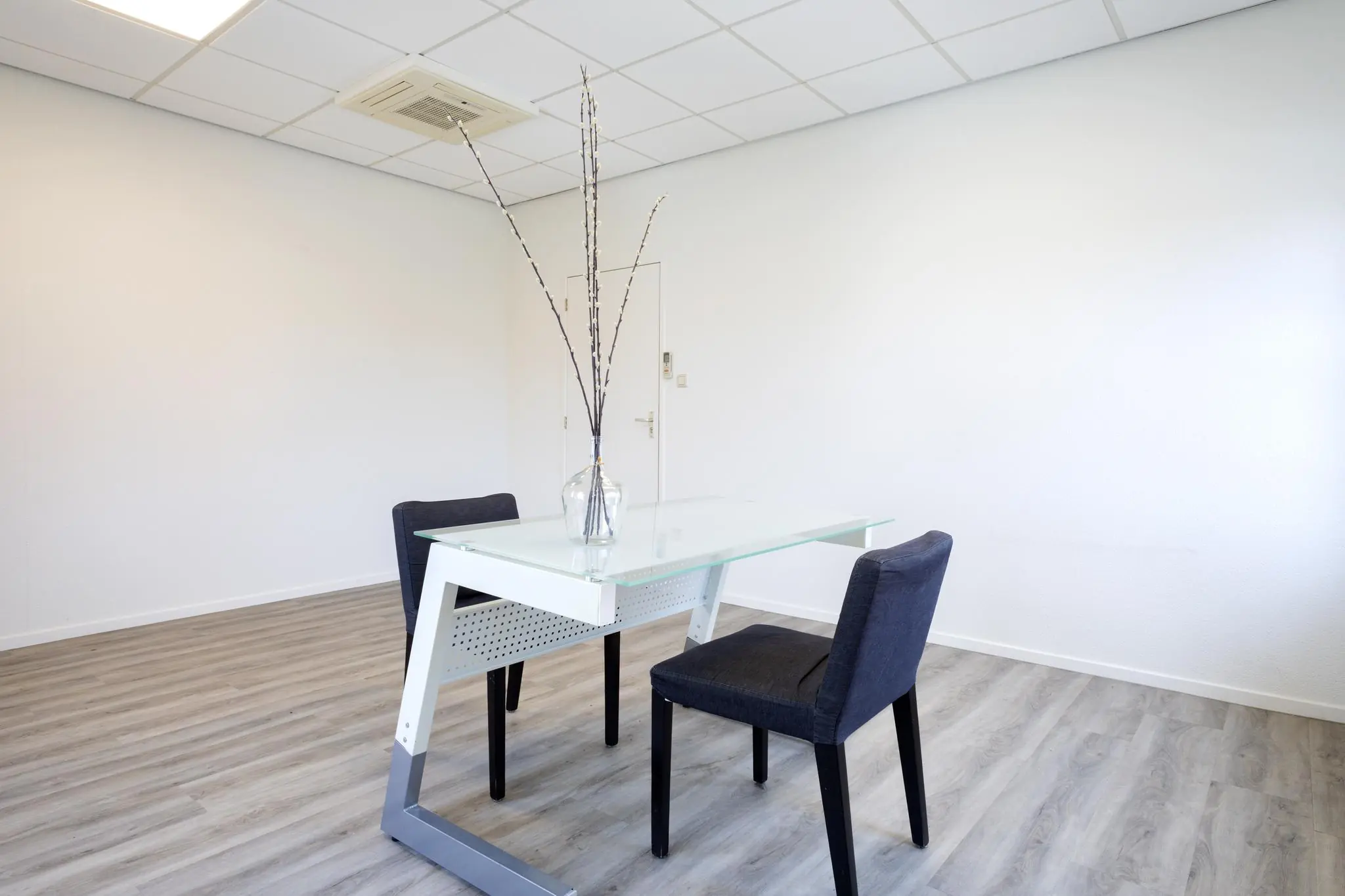 Minimalistisch kantoor met glazen bureau, twee stoelen en een vaas met takken in een lichte ruimte.