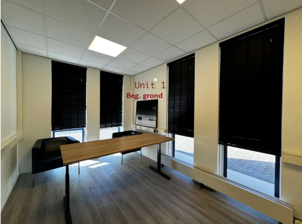 Kantoorruimte met houten bureau, twee zwarte stoelen en meerdere ramen met zwarte jaloezieën in Unit 1 op de begane grond van Bergveste.
