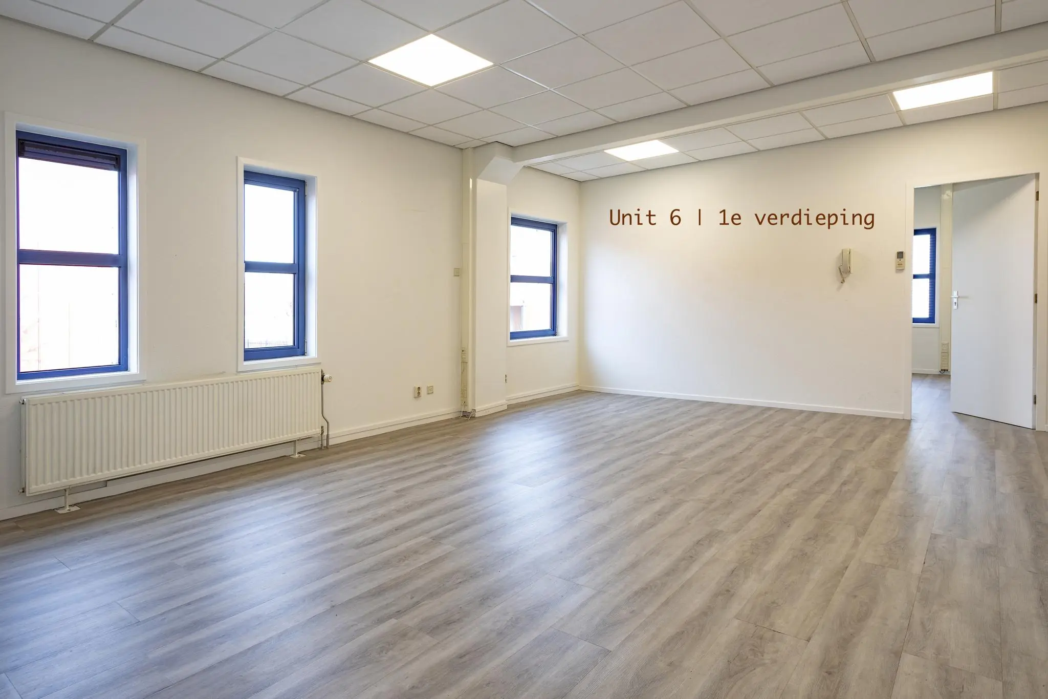 Lichte kantoorruimte met drie ramen, laminaatvloer en de tekst "Unit 6 | 1e verdieping" op de muur.