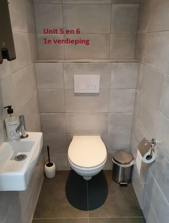 Toiletruimte met hangtoilet, fonteintje en opschrift "Unit 5 en 6 1e verdieping" op de betegelde achterwand.