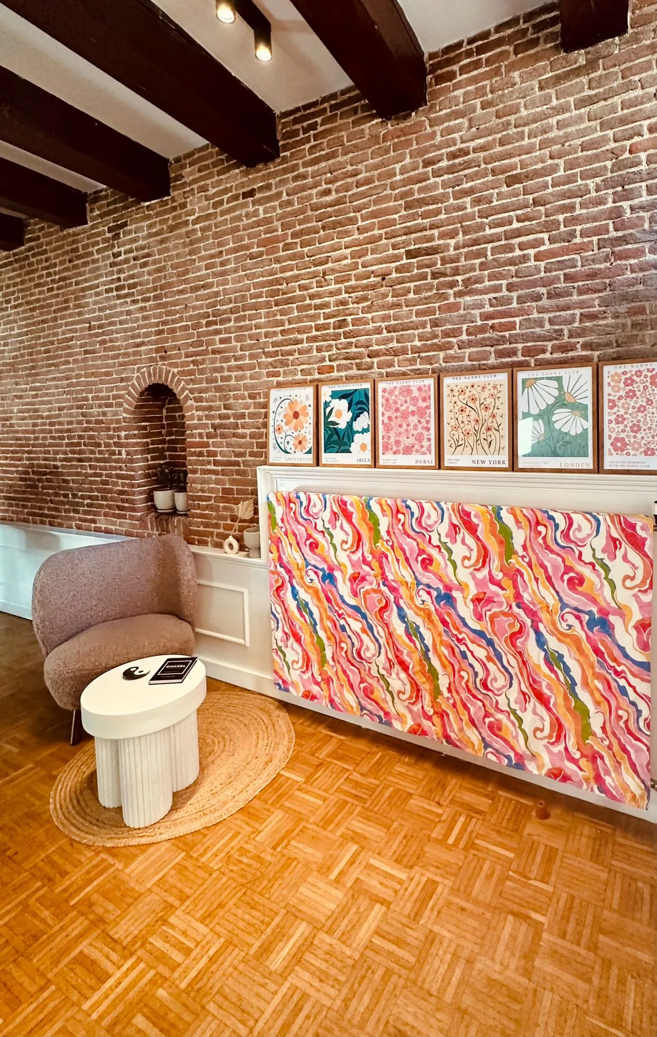 Stijlvol interieur met een kunstzinnige, kleurrijke wanddecoratie en posters van wereldsteden tegen een bakstenen muur.