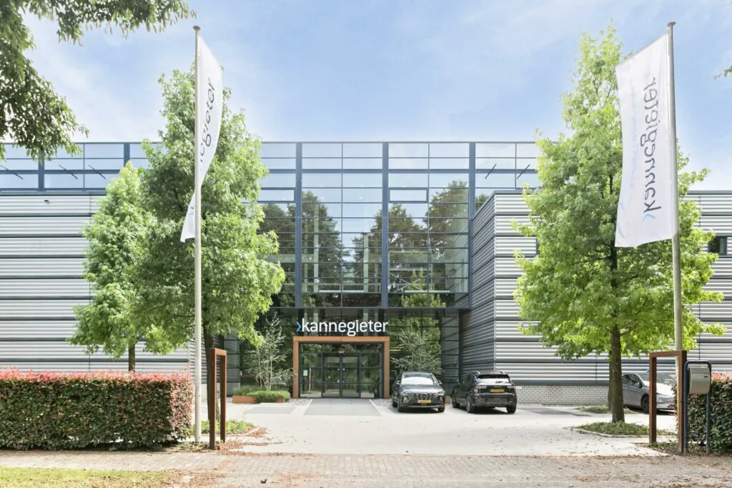 Vooraanzicht van het moderne kantoorpand van Kannegieter met glazen gevel, vlaggen en geparkeerde auto's.