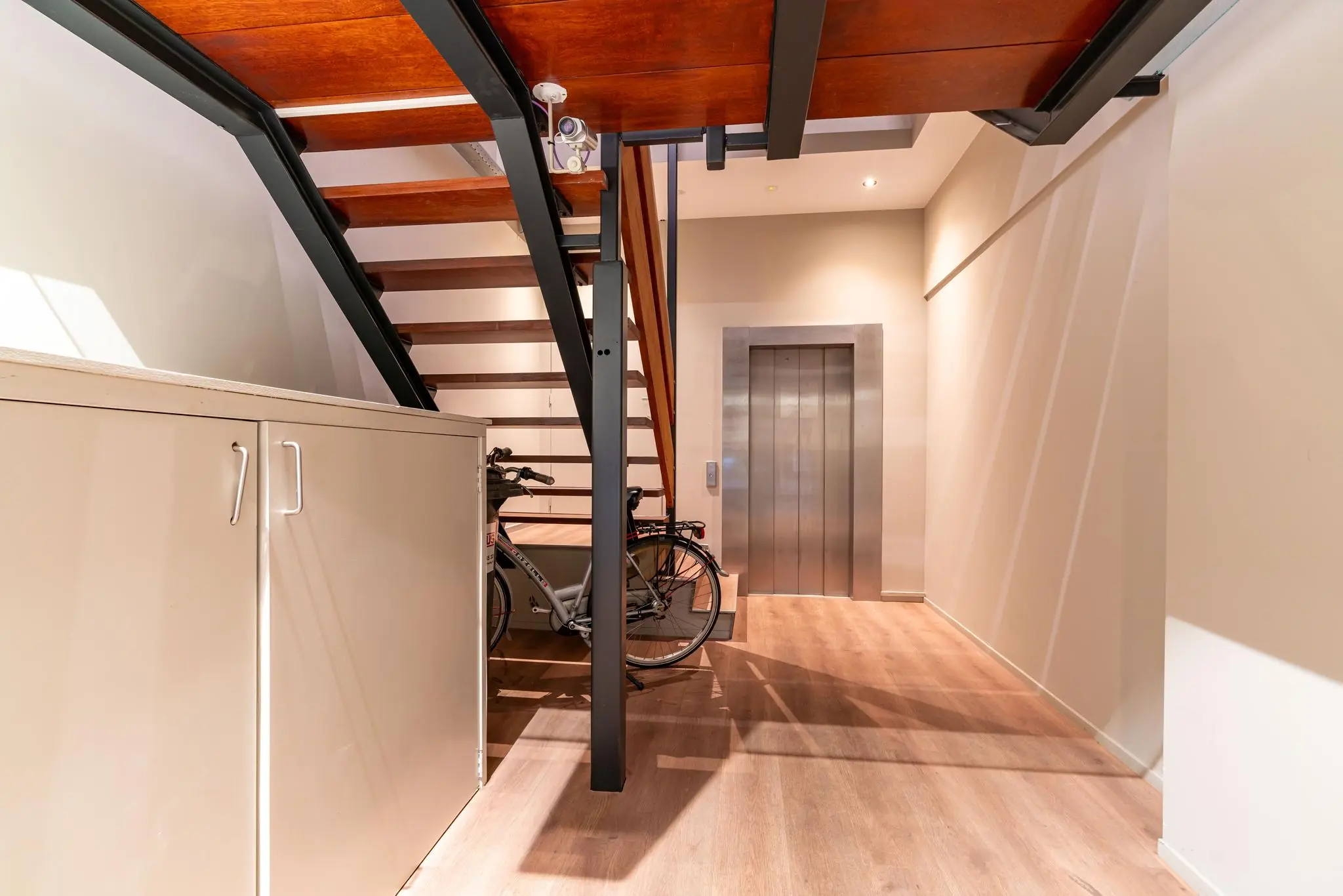 Moderne hal met houten trap, metalen liftdeur, een fiets en een opbergkast.