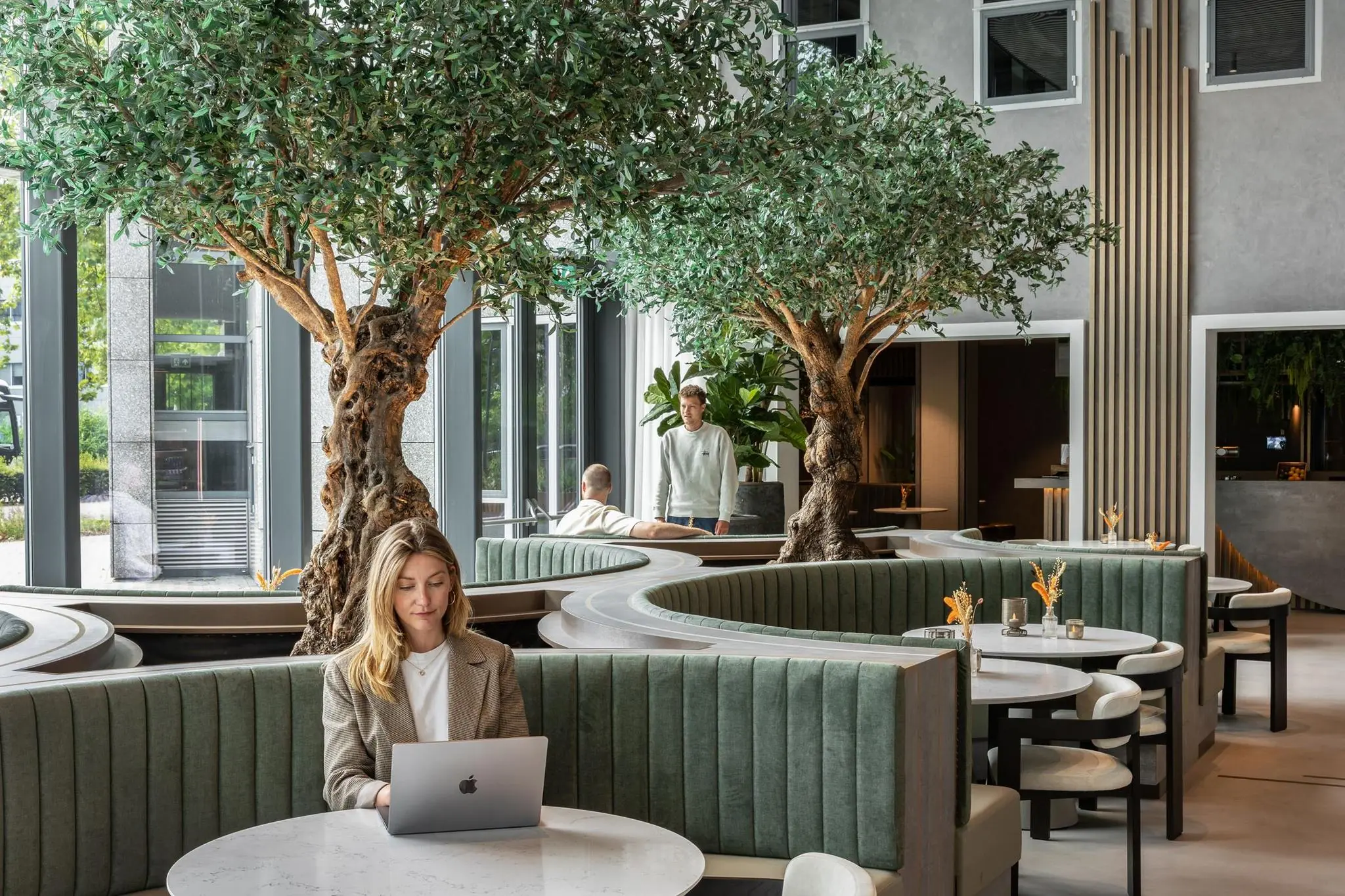 Vrouw werkt op laptop in modern en groen ingericht restaurant Diemerhof met ronde zitbanken en grote indoor bomen.