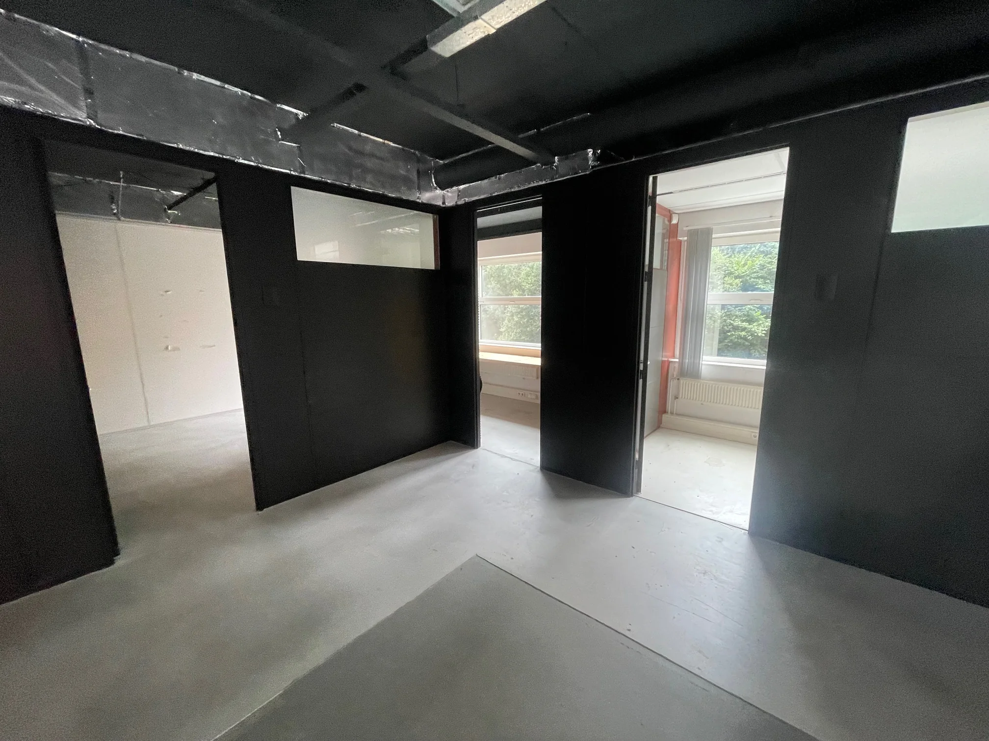 Interieur van een modern, industrieel ogend kantoorpand met zwarte wanden, zichtbare leidingen en grote ramen met uitzicht op groen.