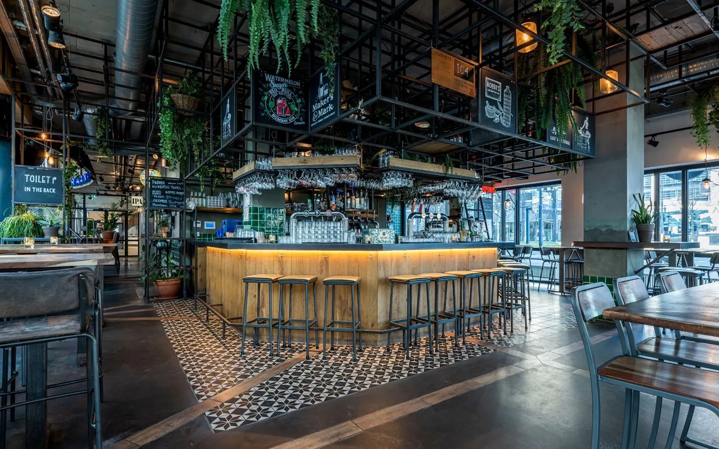 Een moderne en sfeervolle bar met houten meubels, hangende planten en een betegelde vloer.
