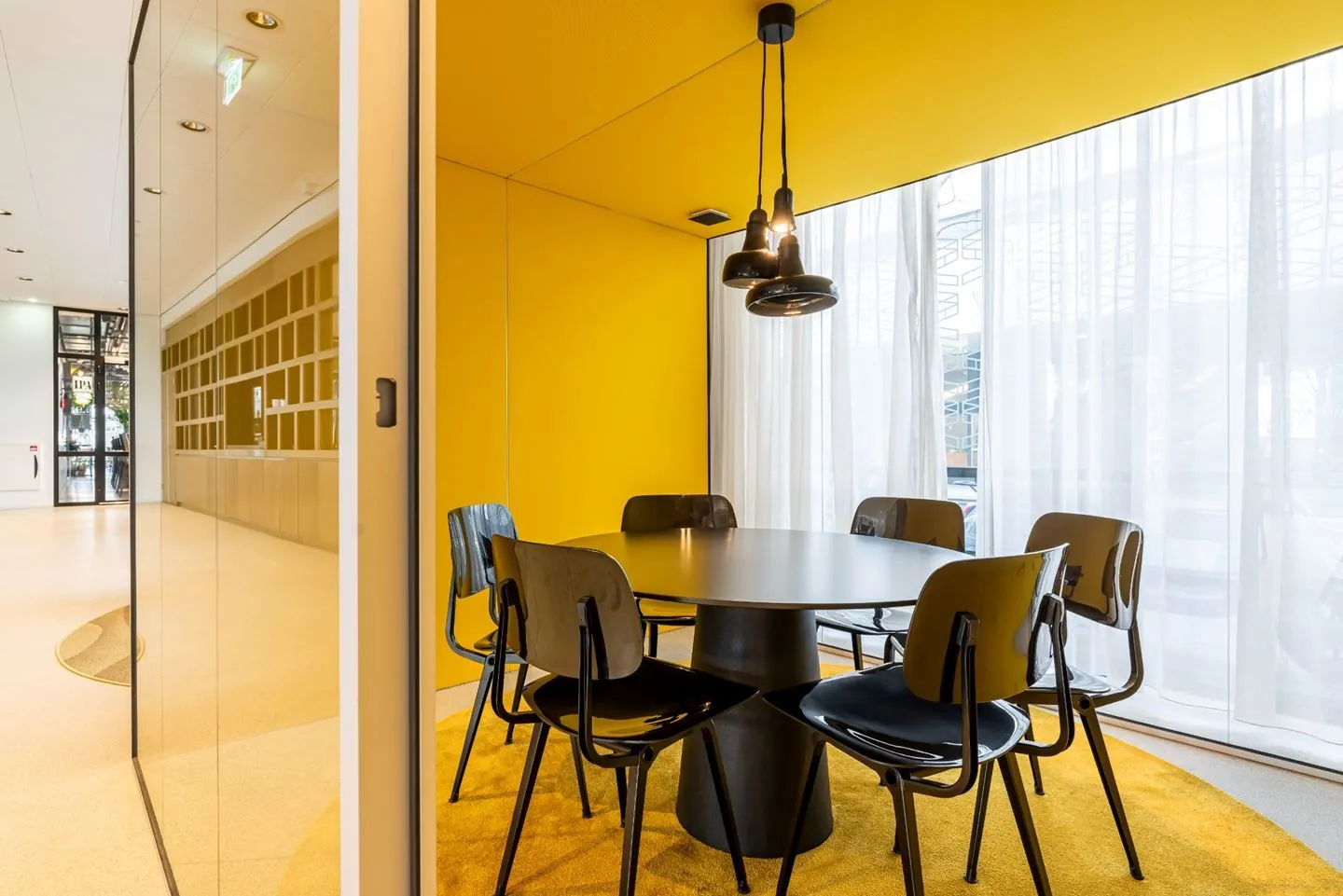 Een moderne vergaderruimte met een ronde tafel, zes stoelen, gele muren en een hanglamp.
