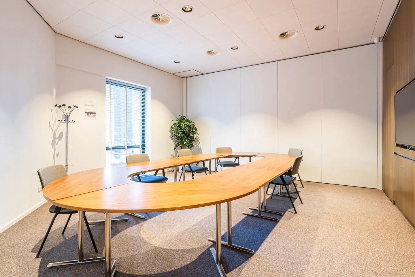 Vergaderzaal met een ovale, houten tafel en stoelen, omgeven door een heldere inrichting en een raam met jaloezieën.