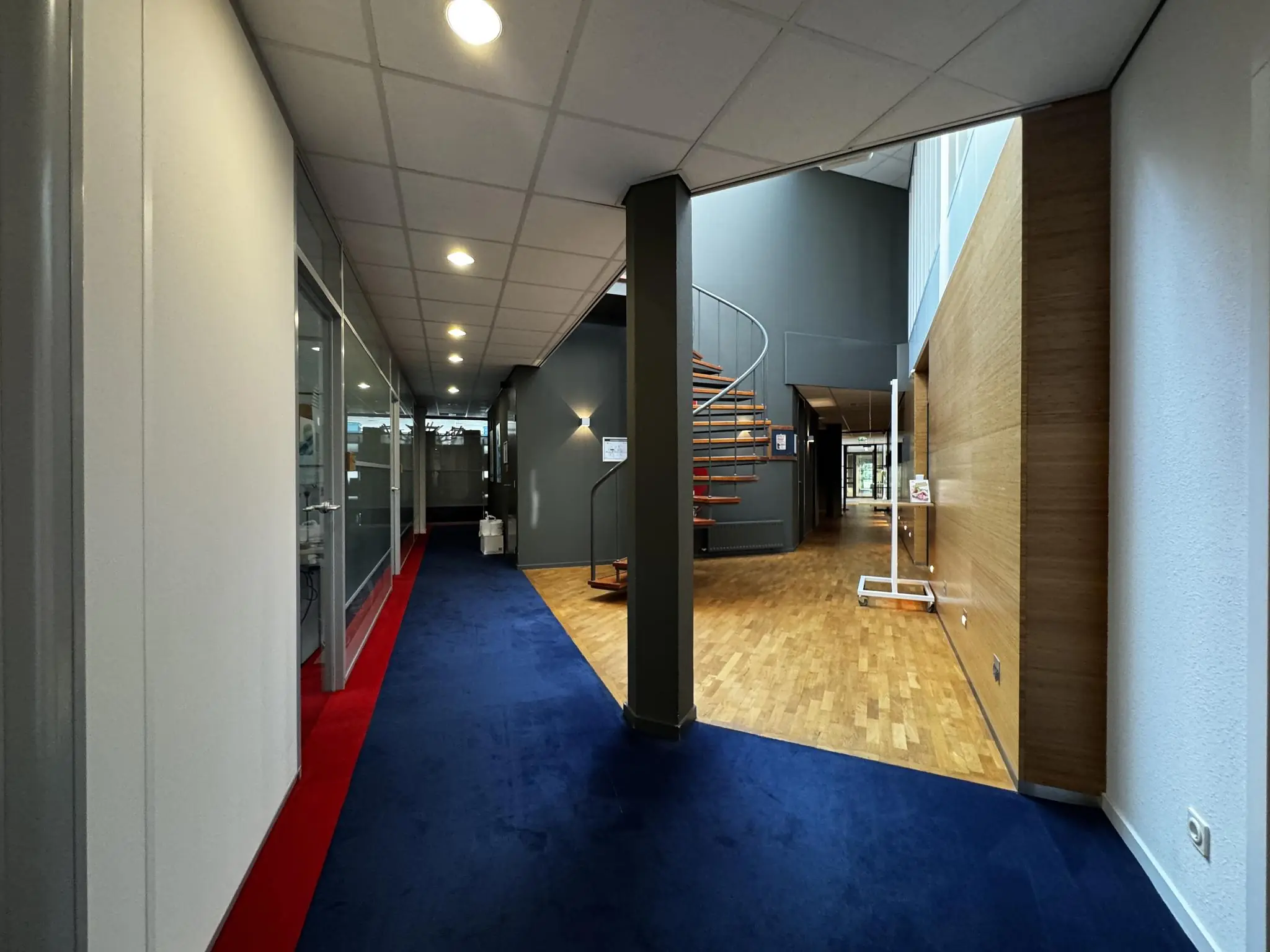 Moderne kantoorruimte met blauwe tapijtvloer, glazen kantoorwanden en een houten wenteltrap in het midden.