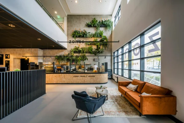 Een modern ingerichte cafélounge met veel natuurlijk licht, een accentmuur met groene planten en een oranje bank.