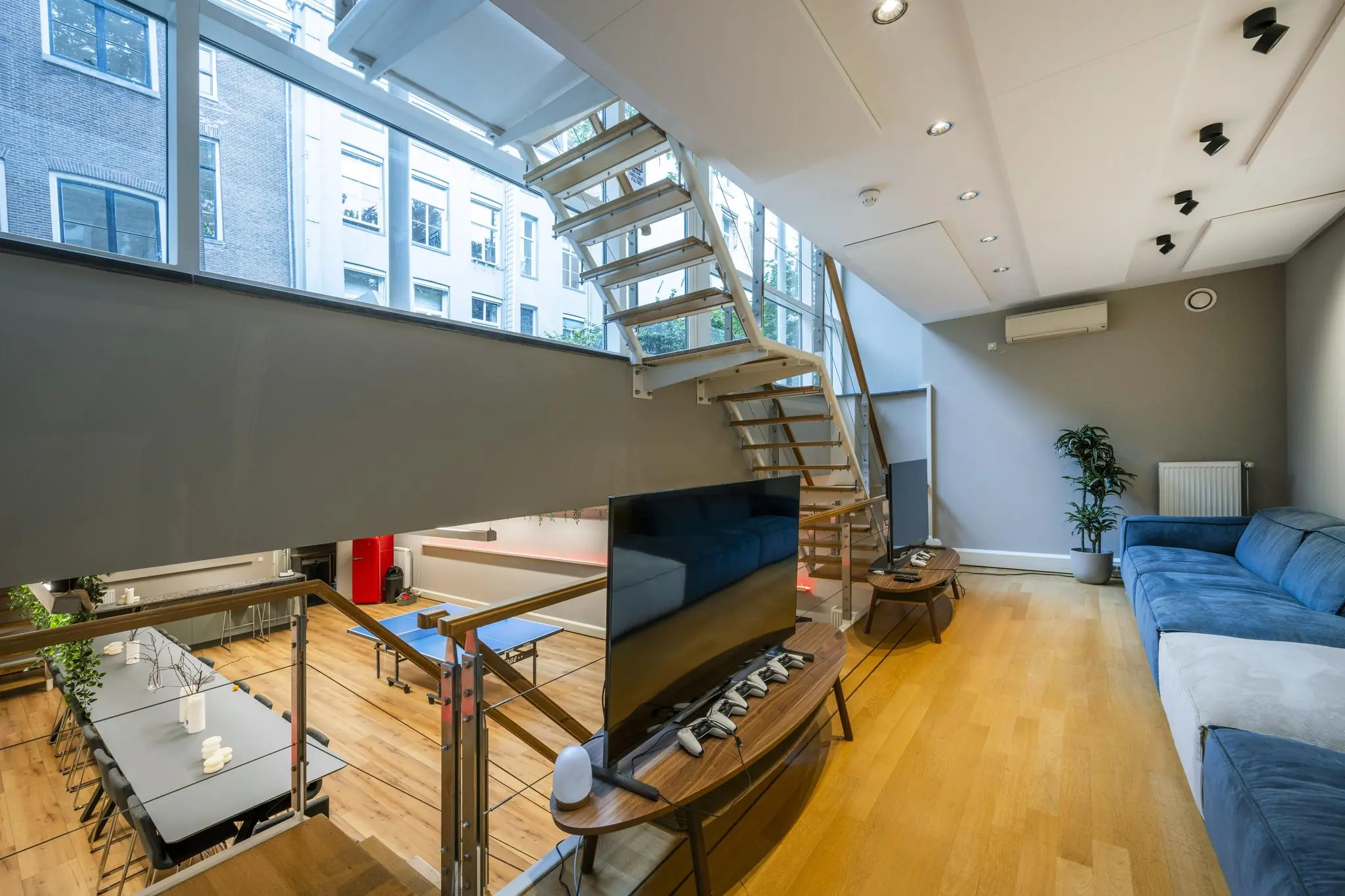 Moderne split-level woonkamer aan de Herengracht met een blauwe bank, open trap en grote ramen die uitzicht bieden op Amsterdamse gevels.