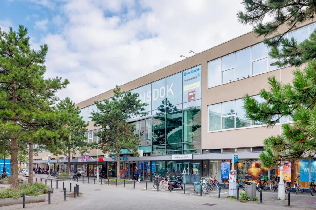 Straatbeeld van de Helmholtzstraat met het gebouw ENSDOK, winkels, fietsen en bomen op de voorgrond.