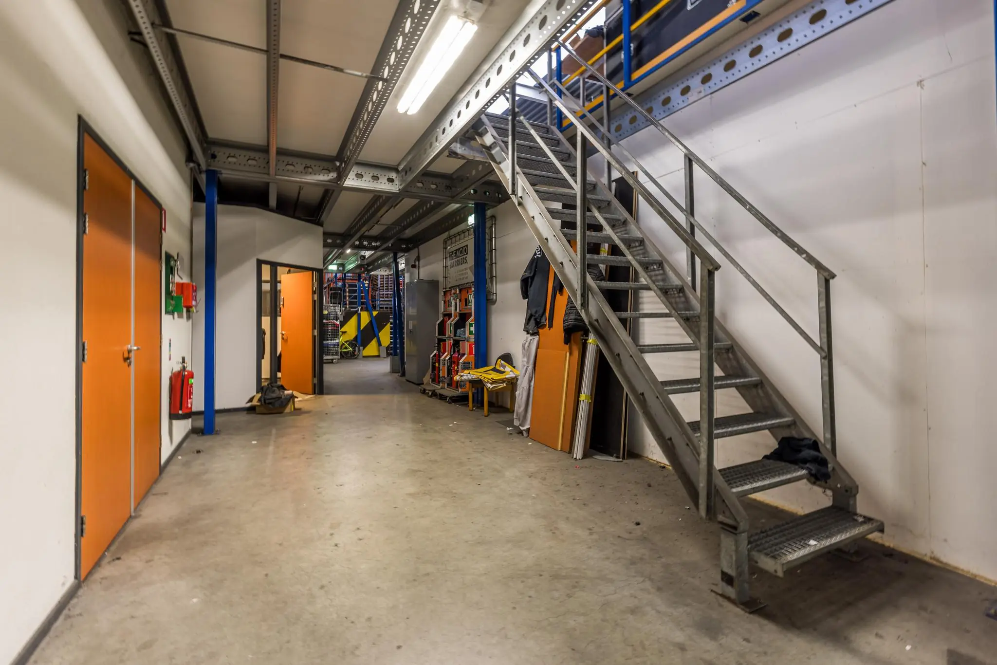 Industriële binnenruimte met metalen trap, oranje deuren en zicht op een magazijn met stellingen op de achtergrond.