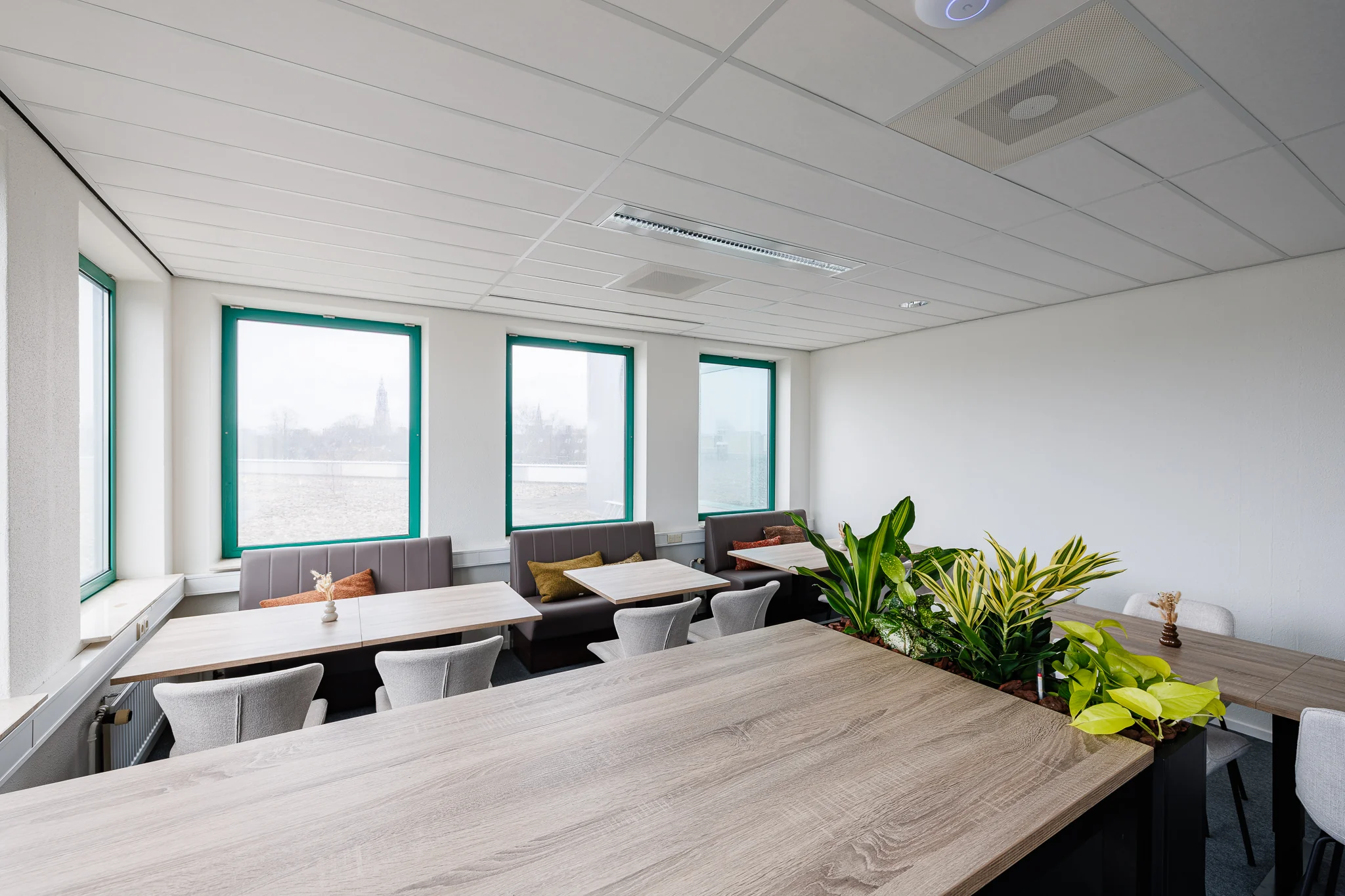 Een moderne vergaderruimte met houten tafels, planten en uitzicht op een stadstoren door grote ramen.