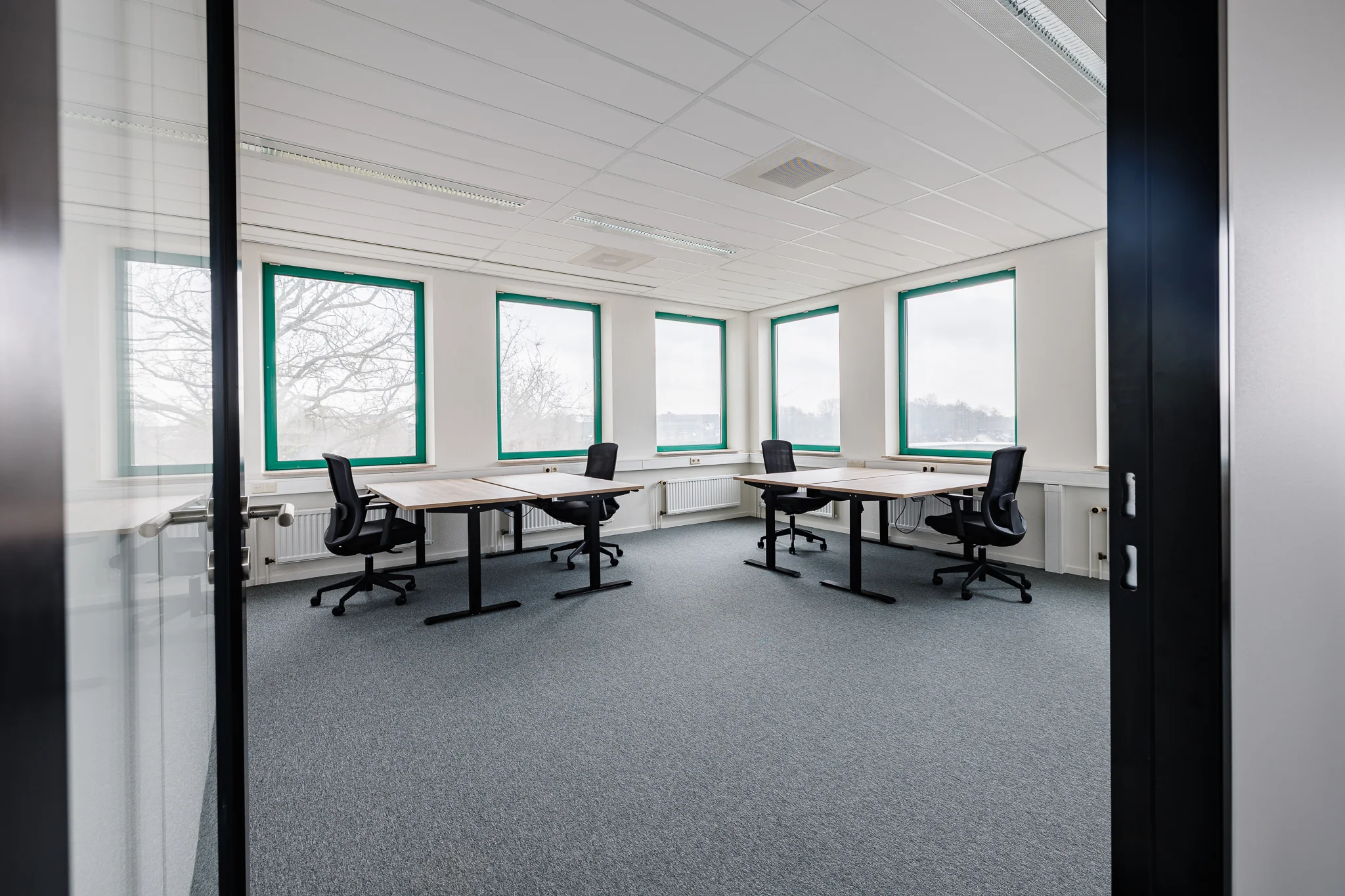 Een moderne, lichte kantoorruimte met vier bureaus en bureaustoelen naast grote ramen.