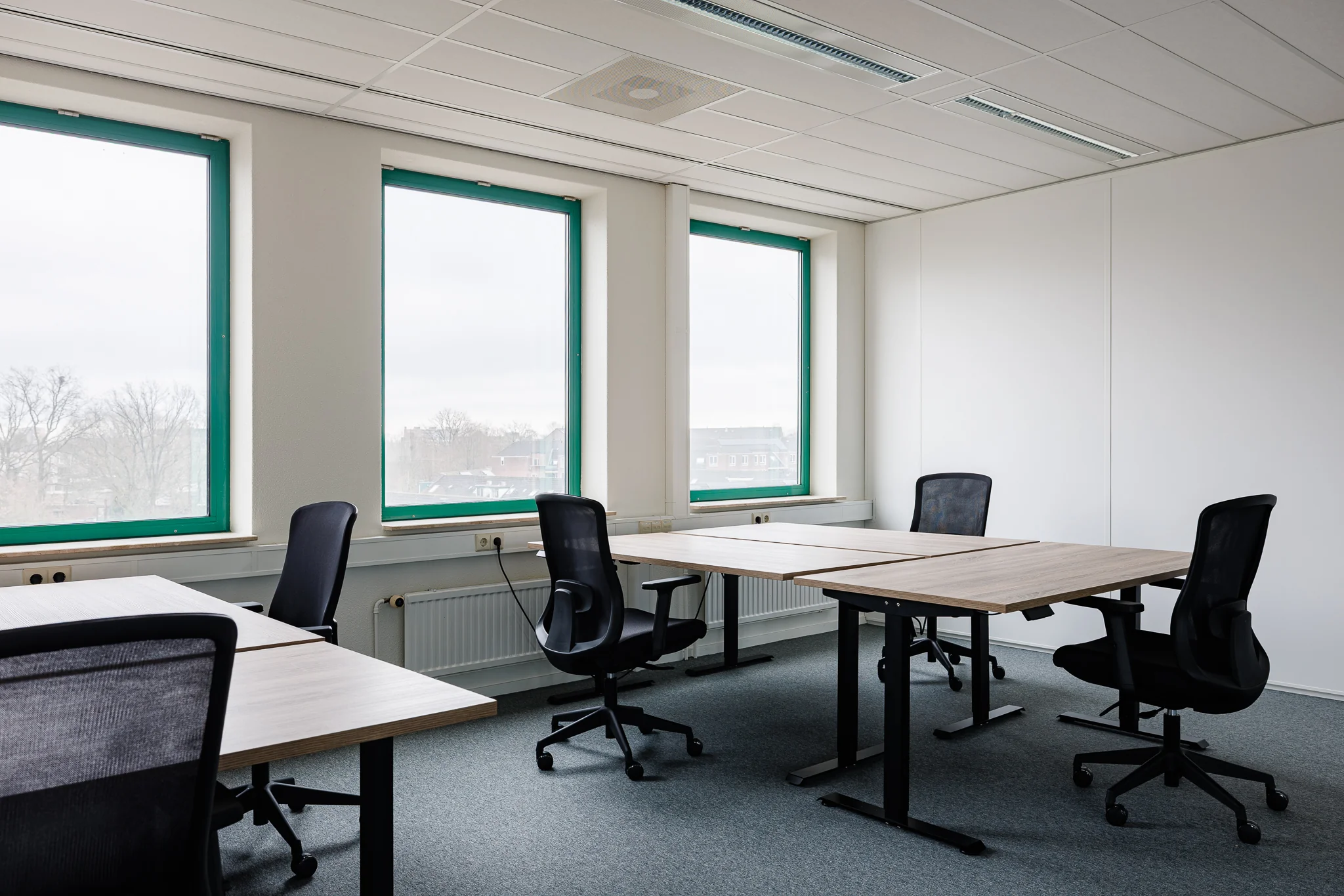 Een moderne, lege kantoorruimte met houten bureaus en zwarte bureaustoelen bij grote ramen.