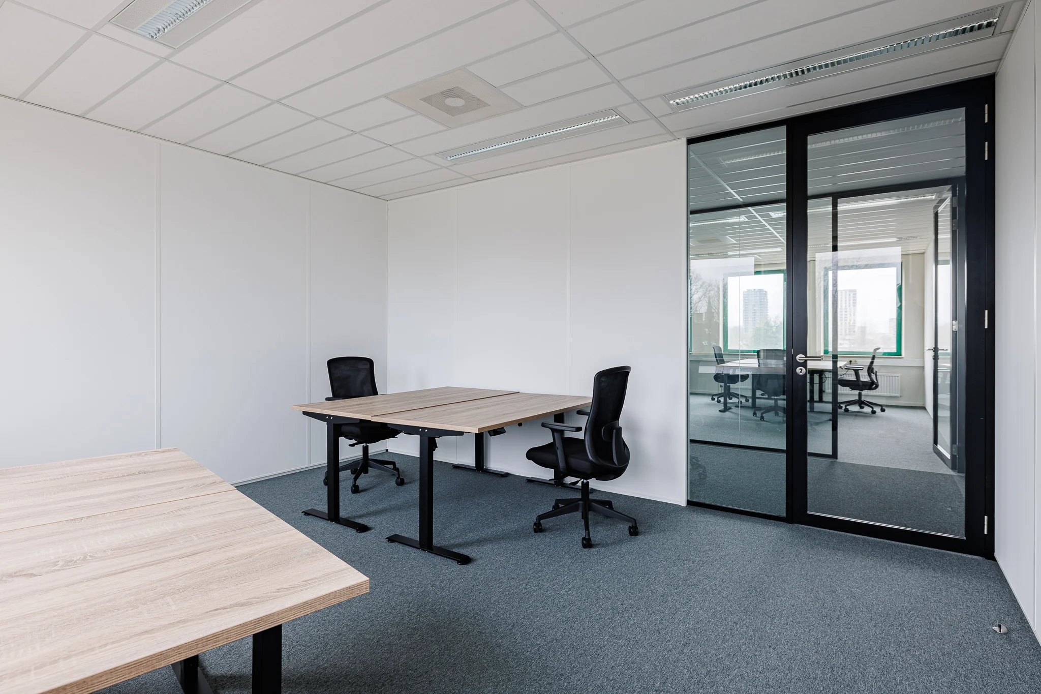 Een modern ingericht kantoor met twee bureaus en zwarte stoelen, gescheiden van een vergaderruimte door een glazen wand.