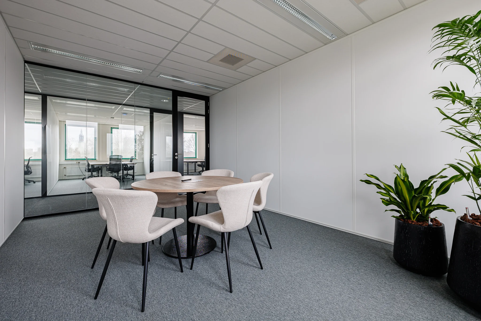 Een moderne vergaderruimte met een ronde houten tafel, vijf beige stoelen en twee grote planten.