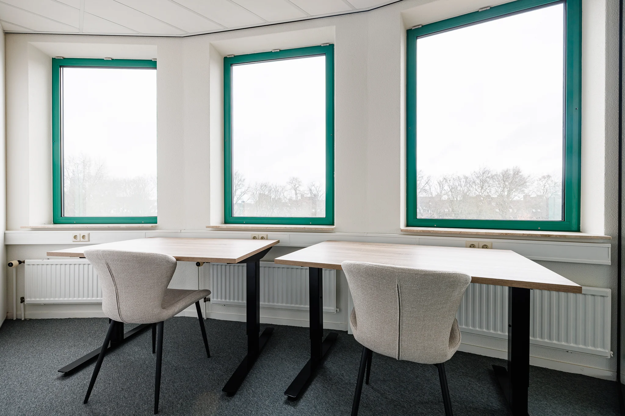 Een moderne kantoorruimte met twee bureaus en stoelen bij grote ramen in een hoekopstelling.