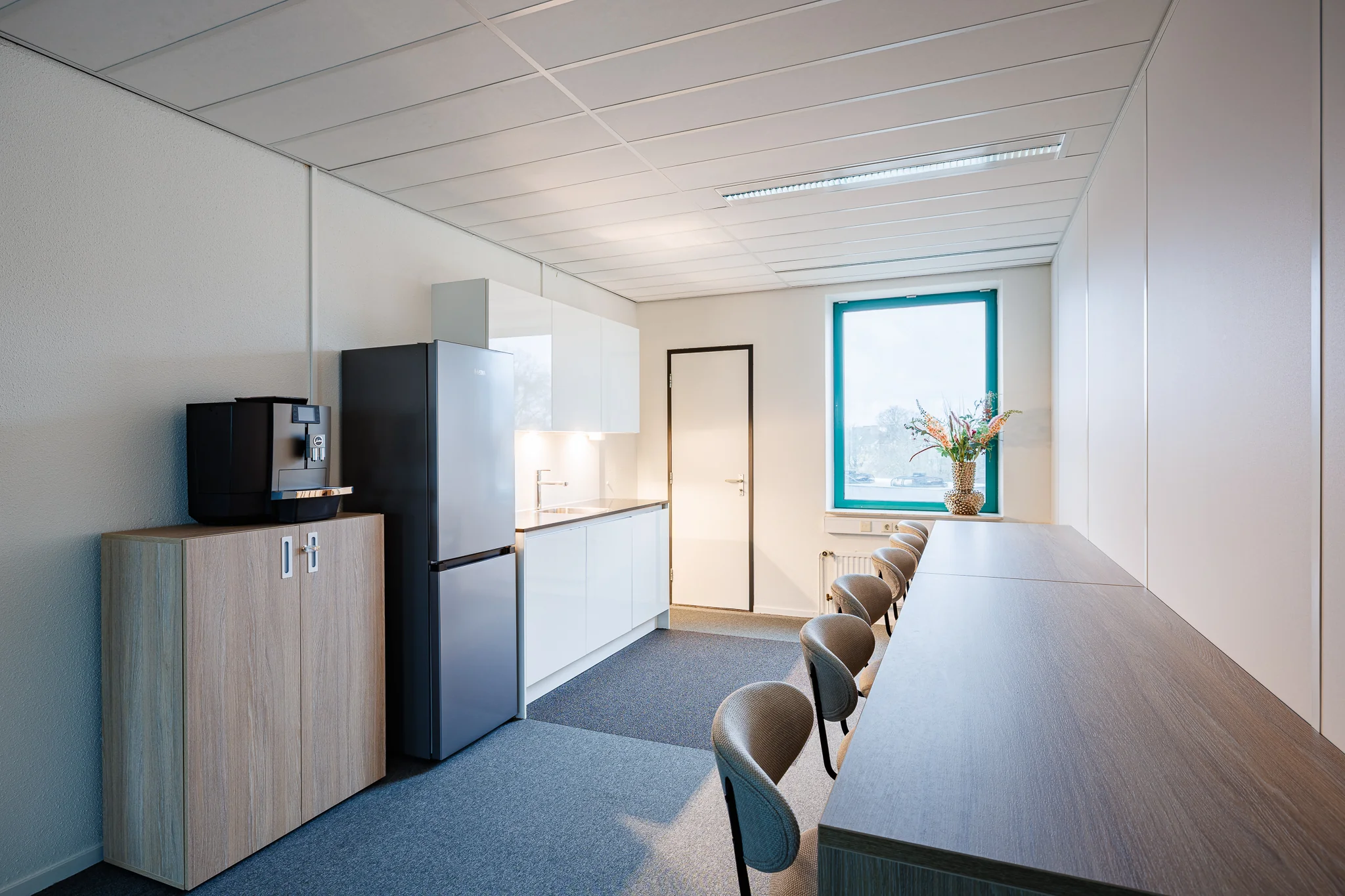 Moderne kantoorruimte in Amersfoort met kitchenette, koffiezetapparaat en lange tafel met stoelen.