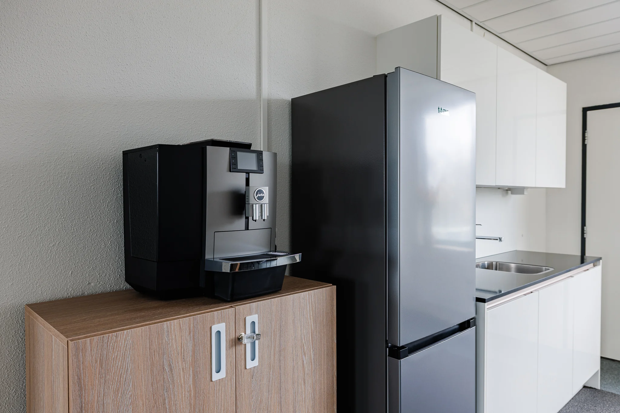 Keuken met een koffiemachine op een houten kast naast een zilverkleurige koelkast en witte kasten.