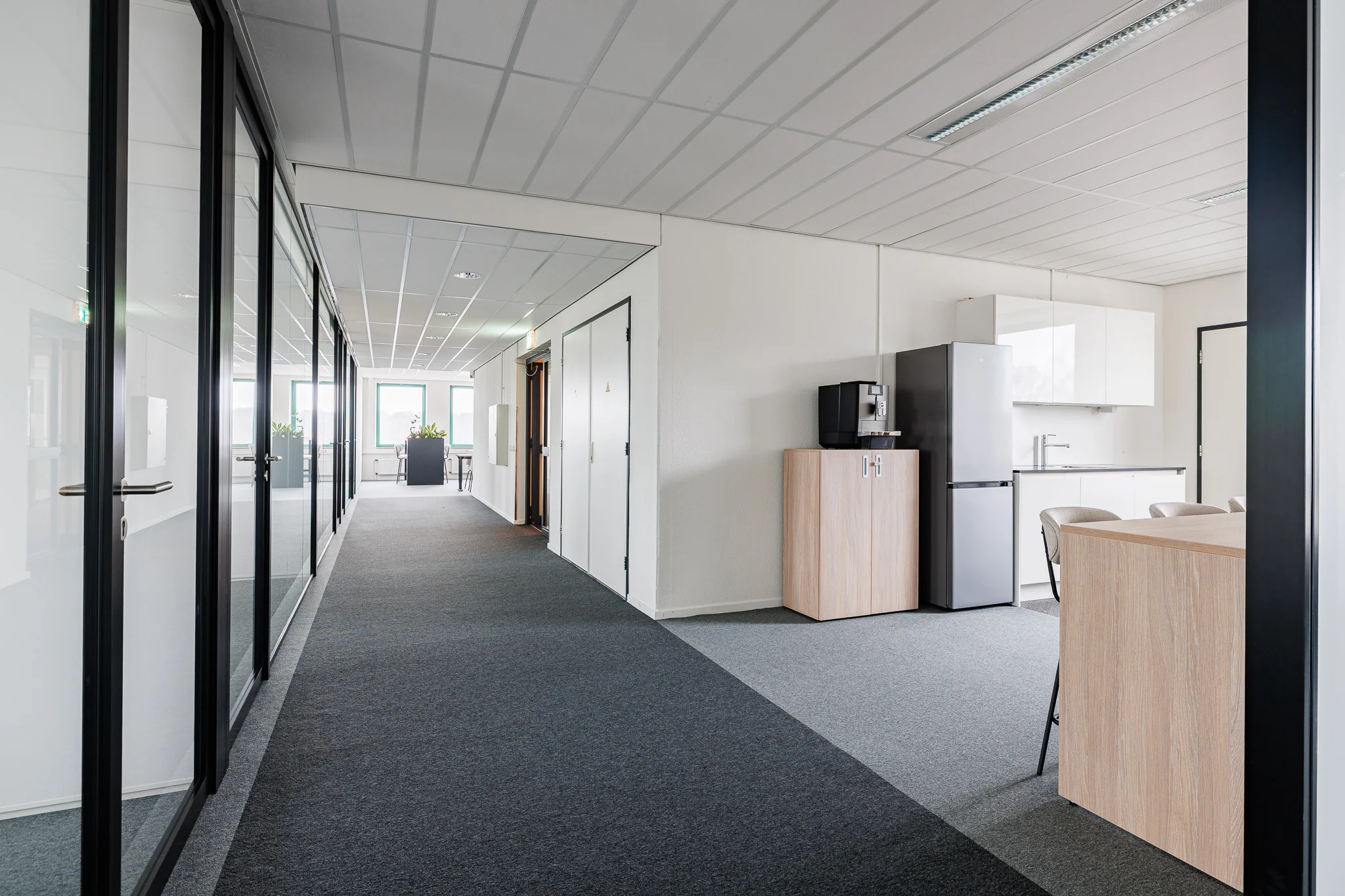 Een modern kantoorinterieur met glazen wanden, waardoor een gang zichtbaar is met zicht op een keukenblok.