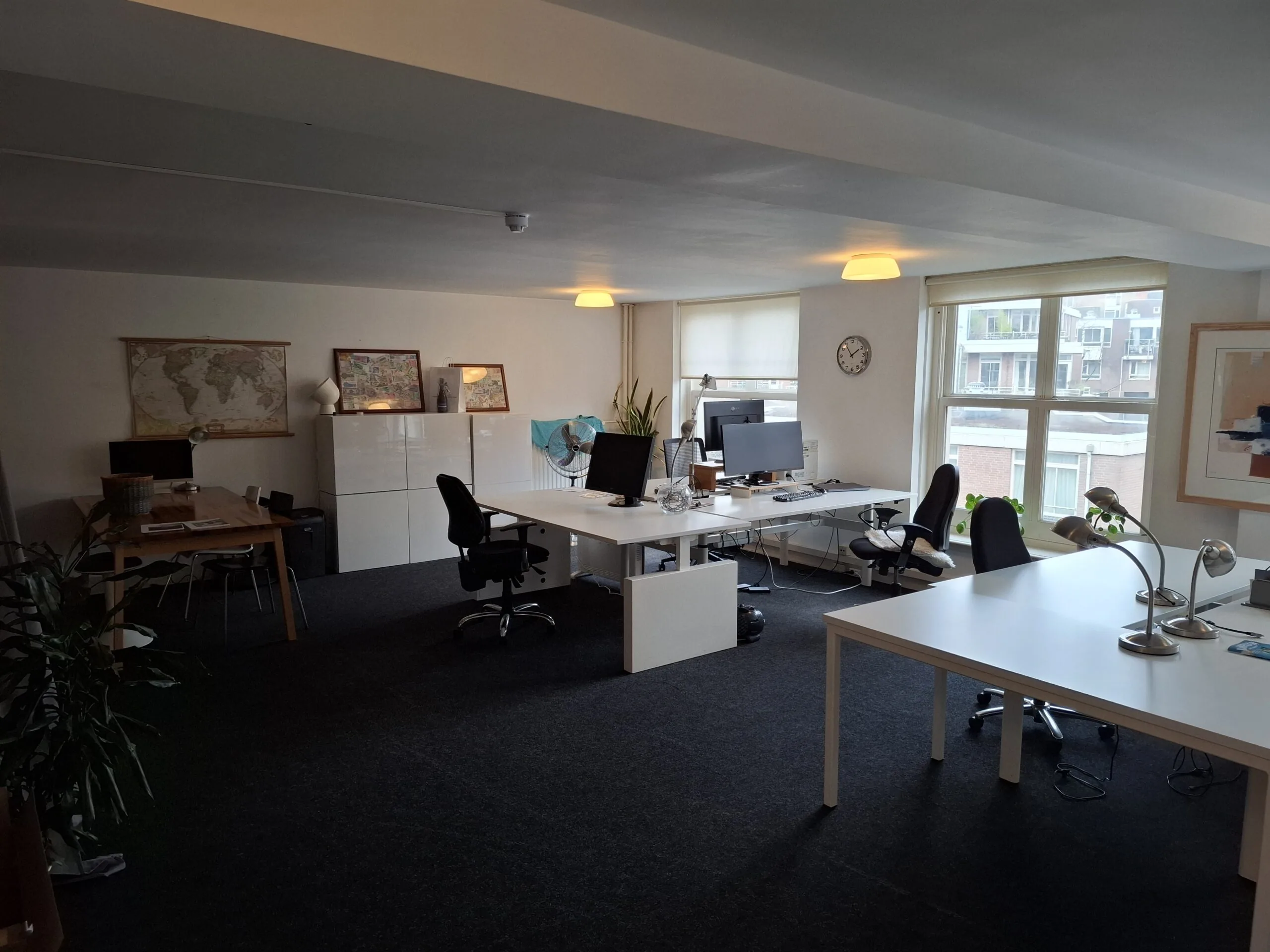 Ruim en licht kantoor met meerdere bureaus, computers, stoelen en grote ramen met uitzicht op een appartementencomplex.