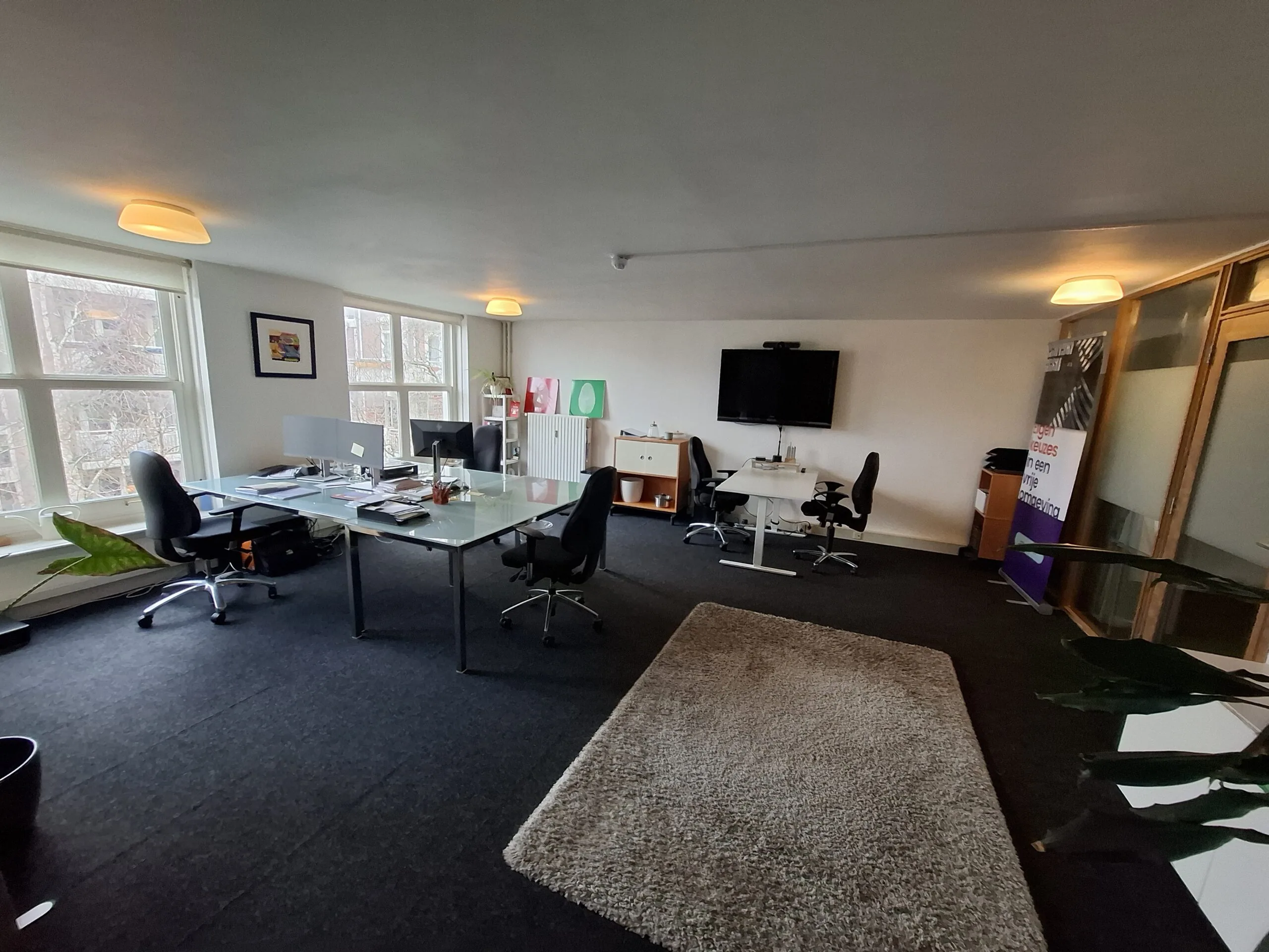 Modern kantoor met bureaus, stoelen, een vergadertafel en een tv-scherm aan de muur.
