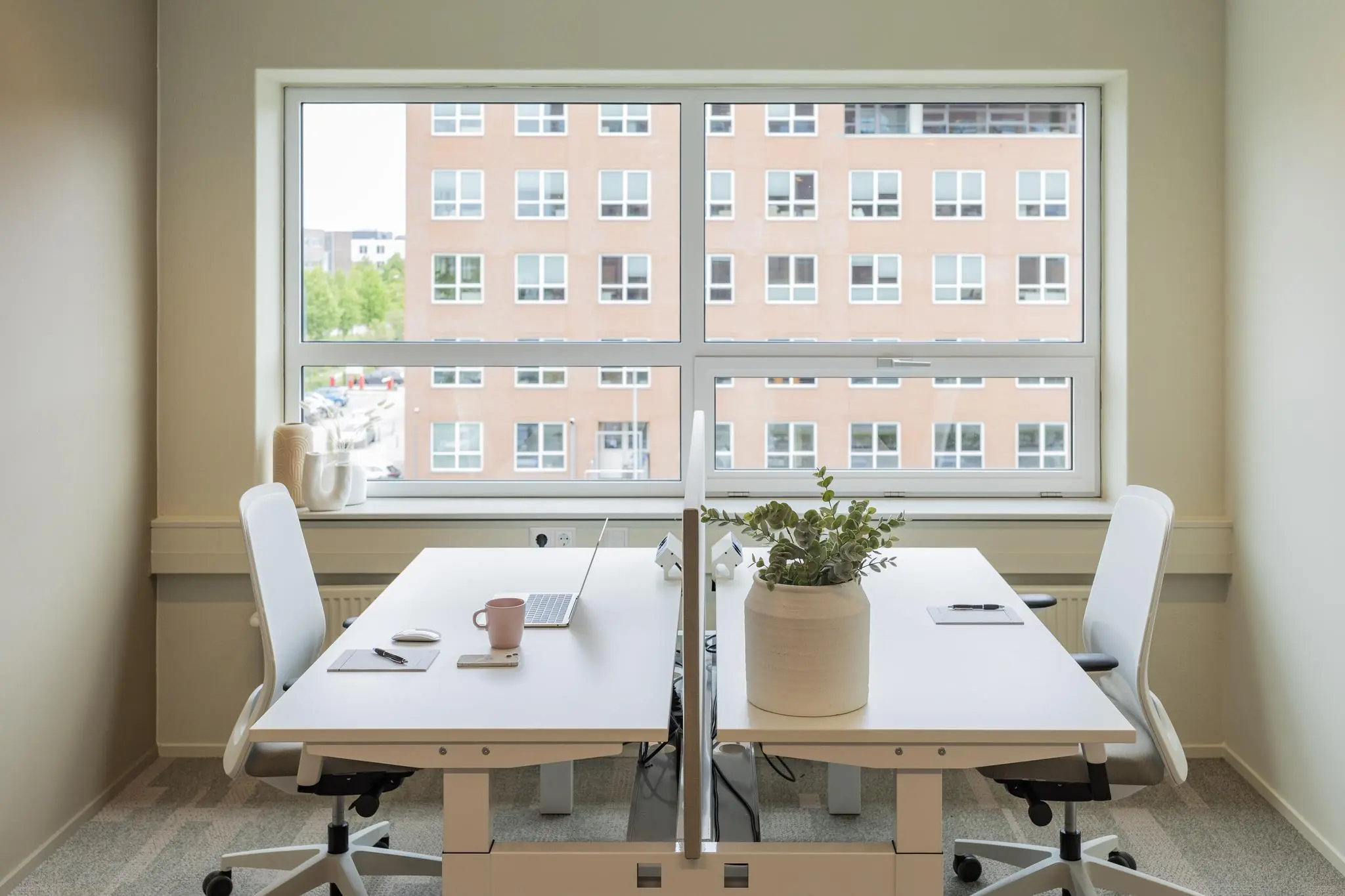 Twee moderne werkplekken met witte bureaus en bureaustoelen bij een groot raam met uitzicht op een bakstenen kantoorgebouw aan de Rijnzathe.
