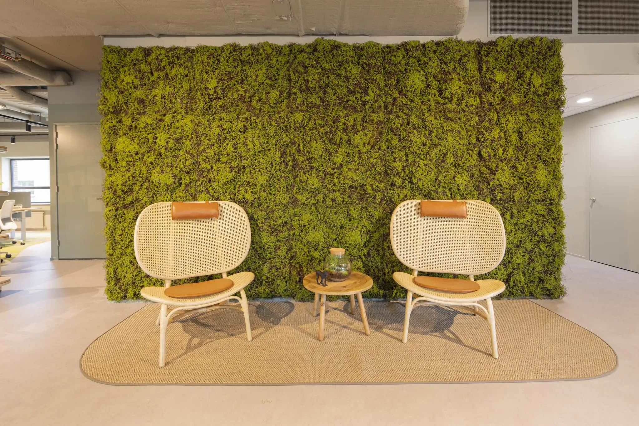 Twee rotan stoelen met leren kussens staan op een vloerkleed voor een groene moswand in een modern kantoor aan de Rijnzathe.