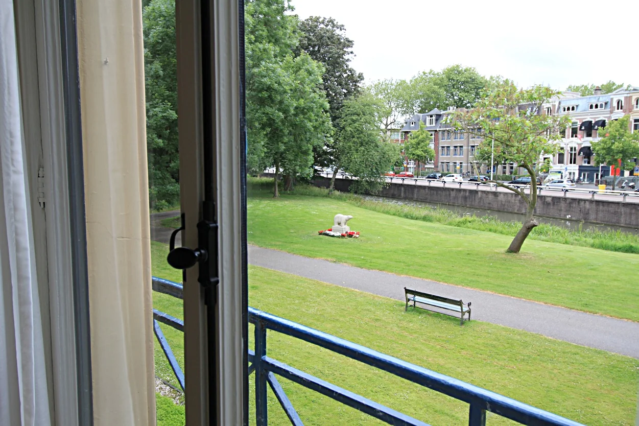 Uitzicht vanuit een open raam op de Museumlaan met een park, een beeldhouwwerk en klassieke herenhuizen op de achtergrond.