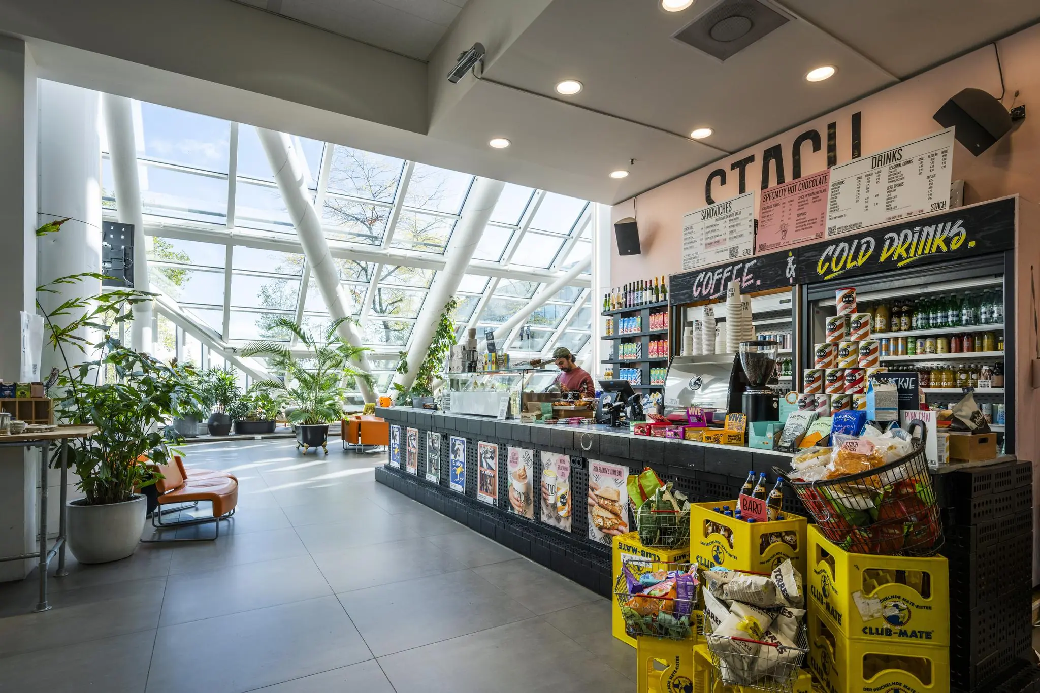 Modern café met grote ramen en veel planten aan de Johan Huizingalaan, met een toonbank vol drankjes en snacks.