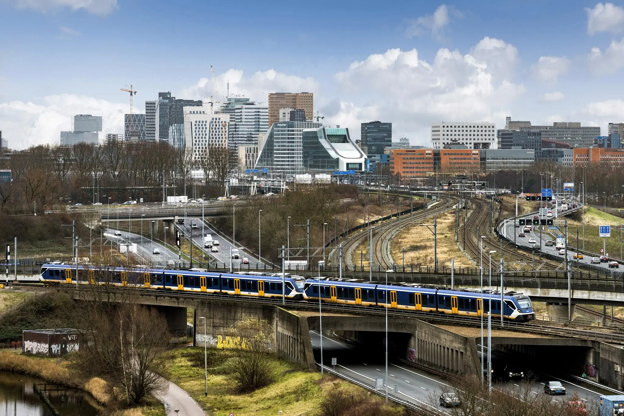 Een NS-trein rijdt over een viaduct op de Johan Huizingalaan met op de achtergrond de skyline van Amsterdam-Zuid.