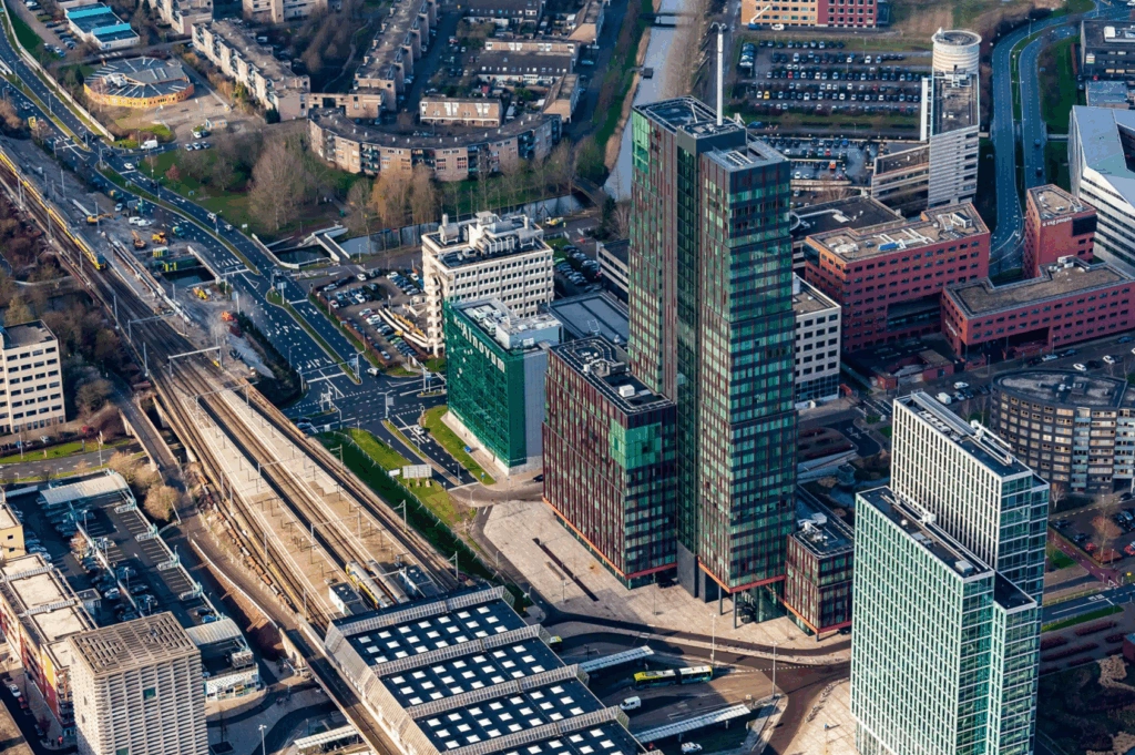 Luchtfoto van de P.J. Oudweg in Almere met moderne kantoorgebouwen, een treinstation en stedelijke infrastructuur.