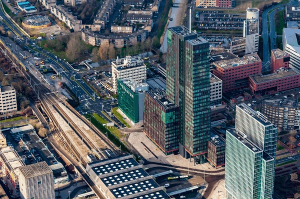 Luchtfoto van de P.J. Oudweg in Almere met moderne kantoorgebouwen, een treinstation en stedelijke infrastructuur.