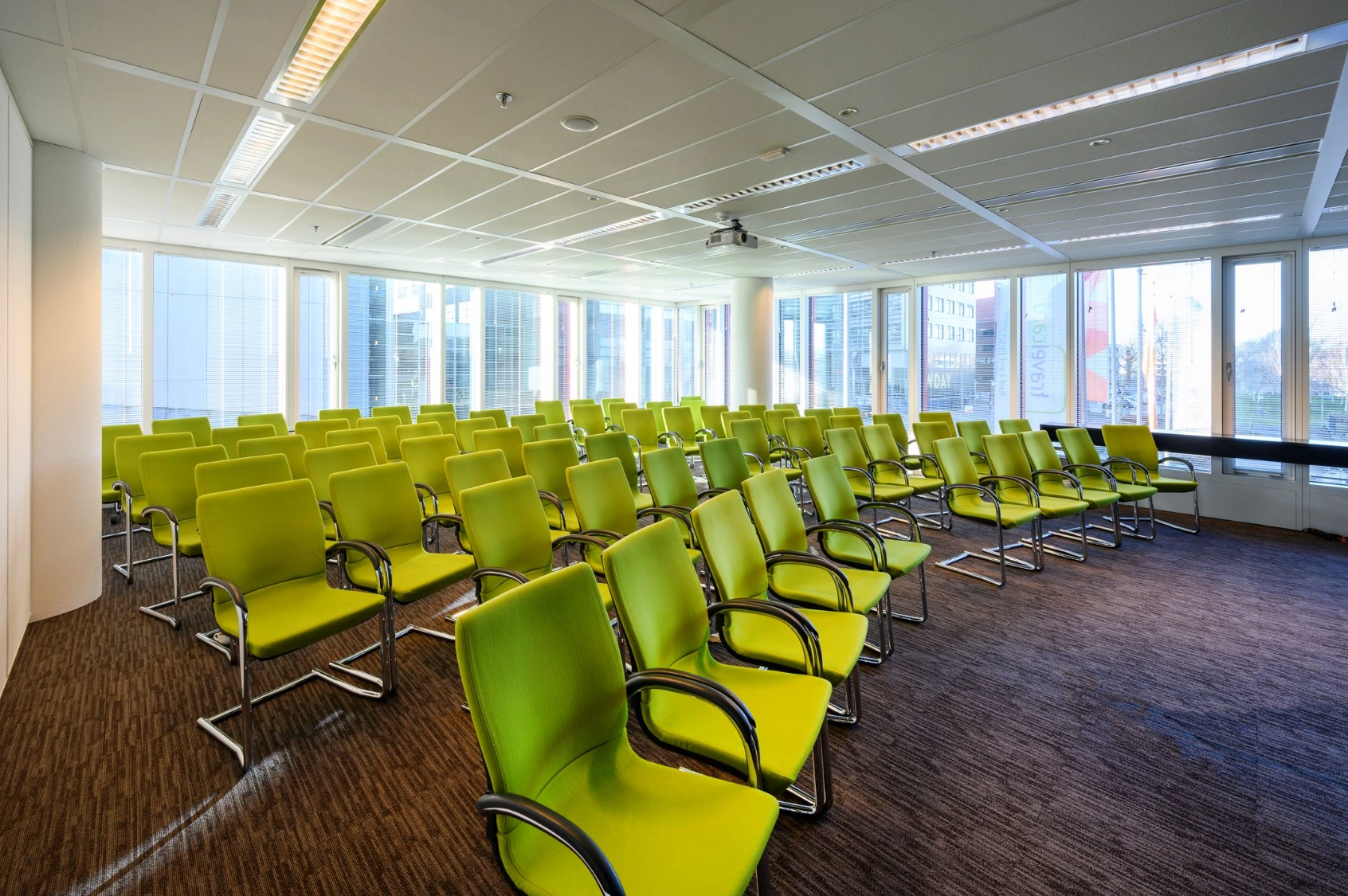 Vergaderruimte aan de P.J. Oudweg met rijen groene stoelen en grote ramen met uitzicht op moderne kantoorpanden.