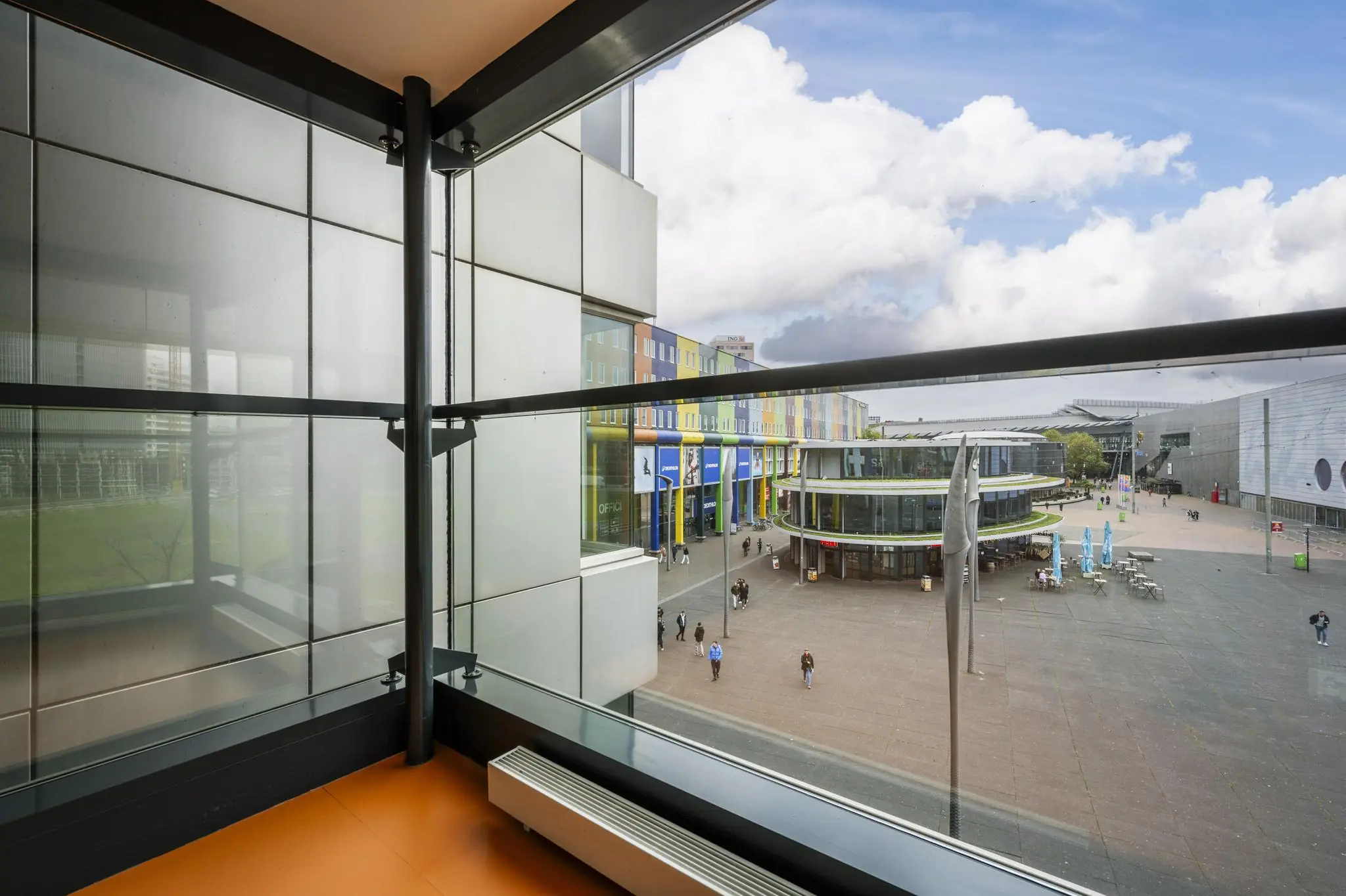 Uitzicht vanaf een balkon op de Johan Cruijff Boulevard met kleurrijke gebouwen, winkels en een modern plein.